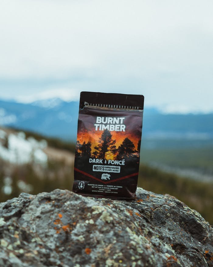 A packet of Burnt Timber coffee sits atop a rocky mountain landscape under a cloudy sky.